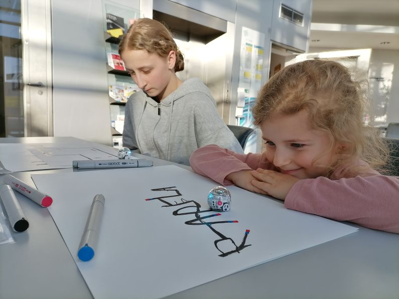 Zwei Mädchen schauen auf Miniroboter, die entlang von Linien fahren.